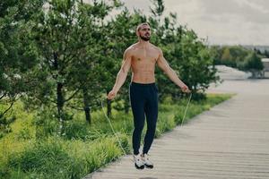 el hombre deportivo con barba espesa hace ejercicios con cuerda de salto, tiene un torso musculoso, se mantiene en buena forma física, posa al aire libre. fitness y concepto saludable. corredor masculino se calienta con equipamiento deportivo foto