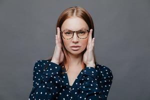 Headshot of serious female teacher keeps hands on head, has self confident facial expression, wears spectacles, polka dot white and black shirt, isolated over grey background. Horizontal shot photo