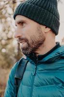 Sideways shot of unshaven man with dark thick beard, wears warm har and anorak, looks pensively aside, poses outside, dreams about something has outdoor stroll in unknown place alone, active lifestyle photo