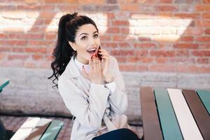 Portrait of surprised beautiful woman with dark pony tail sitting at cafe chatting with her best friend over smartphone gossiping having shocked look after hearing interesting and intriguing news photo