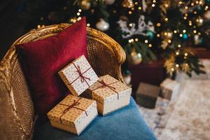 Three wrapped present boxes on armchair near Christmas fir tree. Design or idea for postcard. New Year composition with decorated fir tree and gifts. Majestic atmosphere. Festive event photo