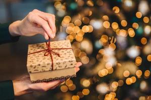 una mujer irreconocible envuelve una caja de regalo sobre un árbol de navidad con luces brillantes y guirnaldas. caja de regalo decorada en manos de mujer. vacaciones y concepto de celebración. foto