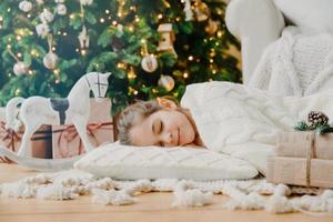 Charming girl sleeps on soft white pillow on floor against decorated New Year tree, has pleasant dreams, surrounded with toy horse and gift boxes. Children, rest and winter holidays concept.