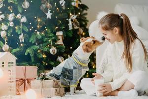 tiempo mágico y ambiente doméstico. feliz niño amistoso y beso de perro, expresa amor y cuidado el uno por el otro, bebe leche fresca, descansa después de decorar el árbol de navidad. niños, mascotas. foto