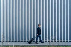 Horizontal panoramic shot of man passenger arrives in own country because of quarantine and pandemic situation in world, walks with suitcase, poses outdoor against metal fence, wears face mask photo