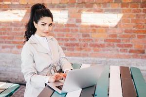 Portrait of cute female model having dark wavy pony tail wearing white elegant coat sitting at desk typing at computer translating article, looking at the camera with serious look posing on brick wall photo