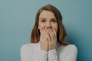 Encantadora mujer joven positiva sorprendida cerrando la boca con ambas manos en emoción foto