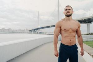 Handsome muscular man with naked torso has outdoor fitness workout looks into distance, has sexy body, dressed in sports trousers stands outside near river bridge. Athletic guy runs in morning
