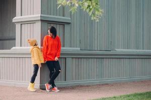 Outdoor shot of friendly woman holds daughters hand, stand at corner of some building, wear warm clothes, enjoy good day and walk in open air, have recreation time. Happy mum and female kid. photo