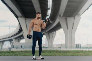 Healthy strong bodybuilder lifts heavy barbells, has workout outdoor, demonstrates his strength, has biceps, stands under bridge maintains good physical shape. Young sportsman works on muscles photo
