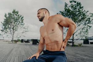 Displeased unshaven European sportsman touches lower back and feels backache after physical training exercises, poses outdoor. Sport exercising injury. Athletic guy suffers pain, needs massage photo