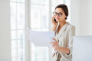 Positive woman in formal clothes, calls to business partner, discusses financial startup project, reads information from papers, wears optical glasses, stands in office, works at international company photo
