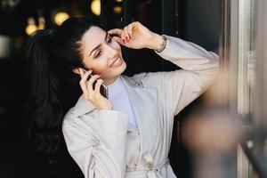 Elegant brunette slim woman with pony tail dressed in white coat having dark eyebrows, eyes, pure skin, nice manicure chatting over mobile phone touching her head with hand looking smiling aside photo