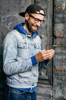 Vertical portrait of hipster guy in denim shirt, cap and eyewear holding modern telephone in his hands having glad expression while communicating with his girlfriend. Lifestyle and technolgy concept photo