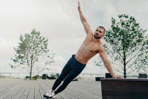 Fit healthy muscular man stands in side plank and raises arm, poses with naked torso, dressed in sport trousers and sneakers, exercises abs, raises abdominal crunch, trains outdoor near beach photo