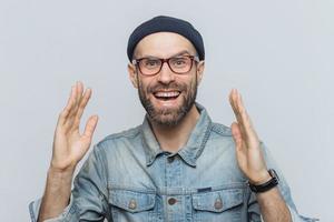 Handsome happy man raises hands with excitment, has overjoyed expression, looks with delighted face, has thick beard and mustache. Glad man in eyewear poses against white background, has fun photo