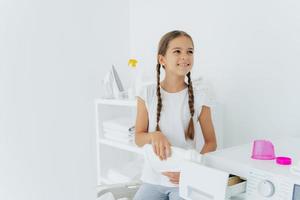 la niña feliz tiene dos coletas que llena la lavadora con detergente líquido, la vierte en la bandeja de la lavadora, se para en el cuarto de lavado con paredes blancas, ayuda a la madre a lavar, usa una camiseta blanca foto