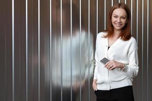 Photo of pleased smiling young European woman dressed in black and white formal clothes, holds smart phone for chatting, reads news from network, smiles happily, stands over wall with copy space