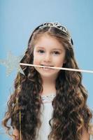 Photo of attractive woman kid with pleasant appearance, has long dark hair, holds magic wand in mouth, poses over blue background, dressed in princess dress. Vertical image. Childhood concept