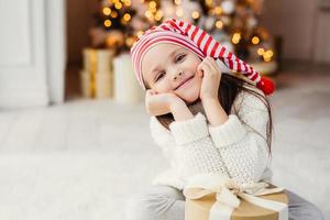 Beautiful small child poses in living room, leans at present gift, has happy expression, glad to recieve surprise from parents, spends holidays in family circle. Merry Christmas and happy New Year photo