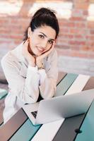 Indoor portrait of young successful Caucasian woman blogger writing a new post for her popular blog using notebook while sitting at desk over brick wall background. Smiling pretty lady with red nails photo