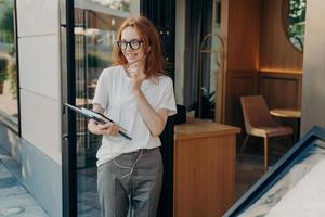 Outdoor shot of glad ginger young woman uses earphones with microphone to communicate photo