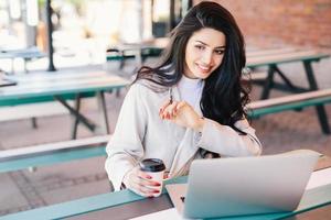 alegre joven morena autónoma que disfruta de conexión inalámbrica gratuita a internet sentada frente a una laptop genérica en un café al aire libre con café para llevar. concepto de relajación, belleza y juventud foto