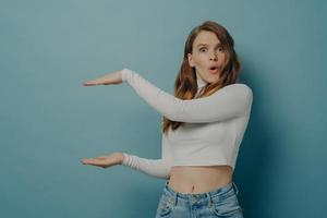 Surprised amazed young woman shows something big in size with hands while standing isolated on blue photo