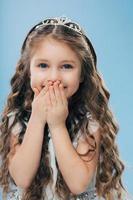 Image of positive beautiful smiling child keeps both hands on mouth, has long thick hair, blue eyes, wears crown, poses against blue background, expresses happiness. Children and satisfaction photo