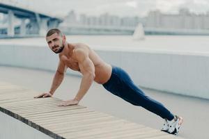 Fit muscular man does plank push up exercise leads healthy lifestyle wears sport trousers and sneakers poses outdoor near bridge. Motivated sportsman pushing hard. Sport, motivation and determination photo