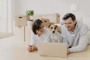 People and new apartment concept. Happy young husband and wife relax on floor, use modern laptop computer for surfing net, pose in their new home or flat, their dog poses near, have little rest photo