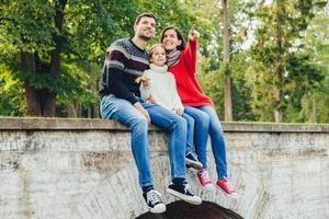 Togetherness and family concept. Friendly father, mother and daughter sit on stone bridge, look from the top, notice beautiful rainbow on sky, indicate on it with fingers, enjoy nature landscapes photo