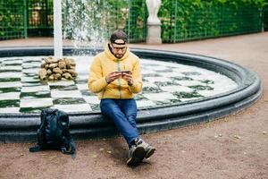 Young attractive bearded man in casual clothes sitting near stone fountain being concentrated into screen of his smartphone watching films while having rest in cozy green park. Man chatting outdoors photo