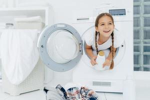 Happy European girl with two plaits, poses inside of wash machine, holds white bottle with liquid powder, has thoughtful expression, pile of dirty clothes in basin, looks somewhere into distance photo