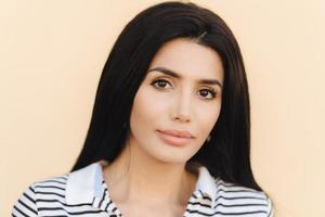 Close up portrait of brunette young female with healthy clean skin, perfect make up, demonstrates her natural beauty, looks directly into camera, dressed in striped dress, isolated on light background photo