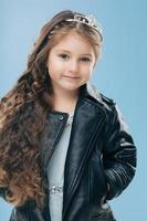 Isolated shot of beautiful Caucasian small child has blue eyes, long dark hair, keeps hands in pockets of leather jacket, isolated over blue background. Children, style, childhood, fashion concept photo