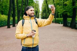 retrato aislado de un hipster con ropa de moda descansando solo en el parque admirando el buen tiempo y el aire fresco. hombre barbudo haciendo selfie en el parque y bebiendo café para llevar de pie en el parque foto