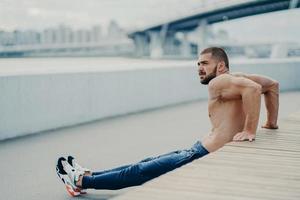 Handsome sportsman trains muscles and makes reverse push ups exercises warms up outdoors before run, concentrated aside, has muscular body, leads active healthy lifestyle, has bare muscular torso photo