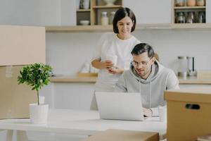 Photo of newlywed couple think about furnishing new bought house, search some ideas in internet on modern laptop computer, drink takeaway coffee and pose in modern kitchen with cardboard boxes