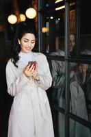 Elegant beautiful young brunette woman with pony tail dressed in white raincoat having pleasant-looking face and perfect red manicure holding smartphone looking into screen with pleasant smile photo