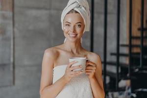 Attractive smiling young woman with makeup, toothy smile, poses bare shoulders, wrapped in towel, drinks hot beverage stands indoor has cheerful expression applies moisturizing cream on face photo