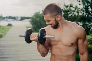 Outdoor shot of strong muscular man with naked torso lifts heavy barbell and trains muscles has workout for keeping healthy fit. Athletic guy demonstrates determination motivates to go in for sport photo