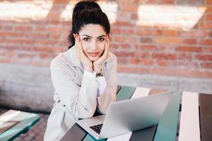 Lovely young brunette woman with charming face and red manicure wearing white coat sitting at table using laptop computer chatting with her family using free internet connection with high speed photo