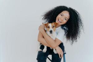 Pleasant looking curly girl tilts head, smiles happily, embraces favourite dog, has good time with pet, wears casual t shirt and jeans, sits on comfortable chair against white background. Relationship photo