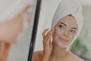 Beautiful woman touches face gently, looks at her reflection in mirror, enjoys softness of skin, stands with wrapped bath towel on head, has minimal makeup and manicure. Beauty treatment concept photo