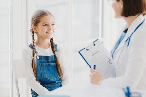 niño feliz escucha atentamente los consejos y prescripciones de los médicos, viene a ver al pediatra en la clínica, tiene dos coletas, usa overoles de mezclilla, habla sobre problemas de salud, tiene un examen de chequeo foto