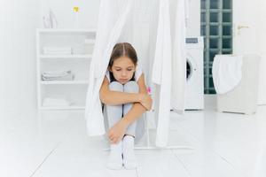Small adorable girl sits on floor, being punished by parents, poses near clothes dryer, focused down with sad expression, washing machine, basket with laundry and console, thinks over something photo