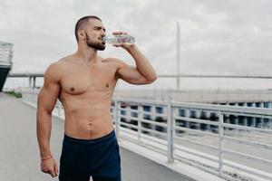 el deportista activo bebe una botella de agua posa con el torso desnudo concentrado en la distancia se siente sediento después de correr al aire libre lleno de energía, lleva un estilo de vida activo. deporte para una vida mejor