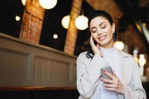Portrait of beautiful female with dark hair tied in pony tail dressed formally listening to music with earphones closing her eyes with relaxation holding cell phone. People, lifestyle, technology