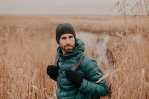 foto de un joven apuesto con rastrojo oscuro, lleva mochila, mira hacia atrás, nota algo, tiene una expresión reflexiva, pasa el tiempo libre al aire libre. otoño. concepto de personas y aventuras